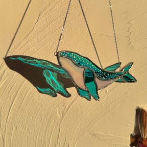 May include: A stained glass whale hanging from a silver chain. The whale is teal blue with white polka dots and a pink belly. The whale is casting a shadow on the wall behind it.