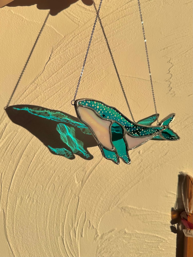 May include: A stained glass whale hanging from a silver chain. The whale is teal blue with white polka dots and a pink belly. The whale is casting a shadow on the wall behind it.