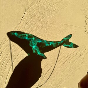 May include: A teal blue whale pendant necklace casting a shadow on a textured beige wall.