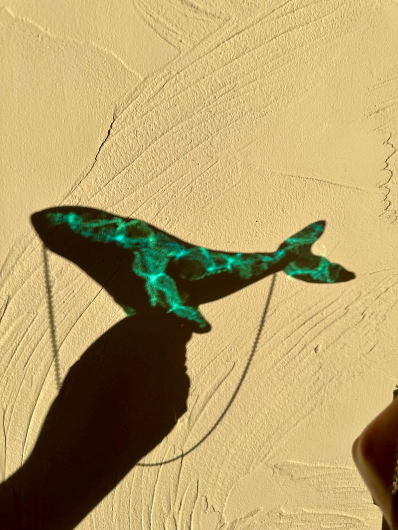 May include: A teal blue whale pendant necklace casting a shadow on a textured beige wall.