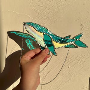 May include: A stained glass whale hanging from a silver chain. The whale is teal blue with white polka dots and iridescent accents.