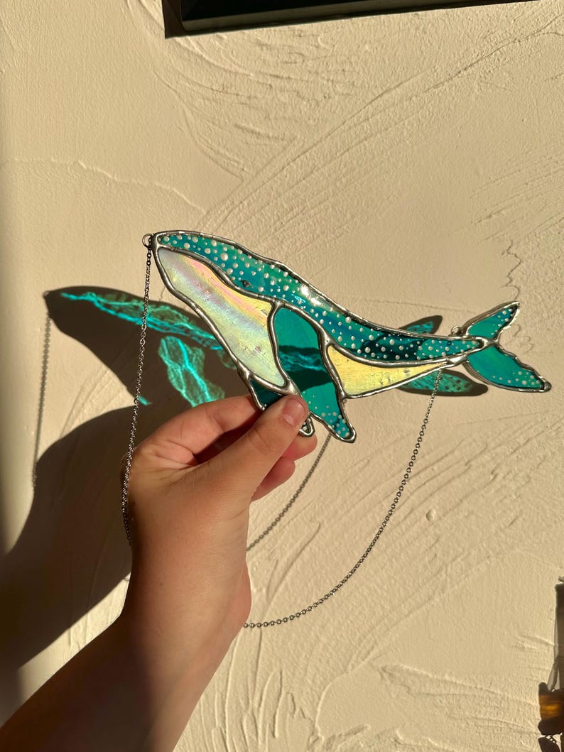 May include: A stained glass whale hanging from a silver chain. The whale is teal blue with white polka dots and iridescent accents.