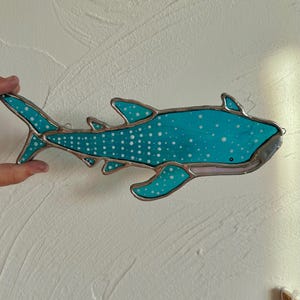 May include: A stained glass whale shark with a turquoise body and white spots. The shark has a silver border and fins. The artwork is hanging on a white wall.