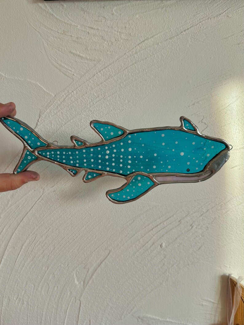 May include: A stained glass whale shark with a turquoise body and white spots. The shark has a silver border and fins. The artwork is hanging on a white wall.