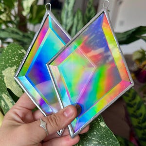 May include: Two diamond-shaped stained glass suncatchers featuring iridescent, holographic surfaces that reflect a spectrum of rainbow colors. Each piece is framed in silver-toned metal solder and includes a small metal loop at the top for hanging.