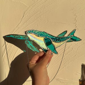 May include: A stained glass whale hanging from a silver chain. The whale is teal blue with white spots and iridescent accents.