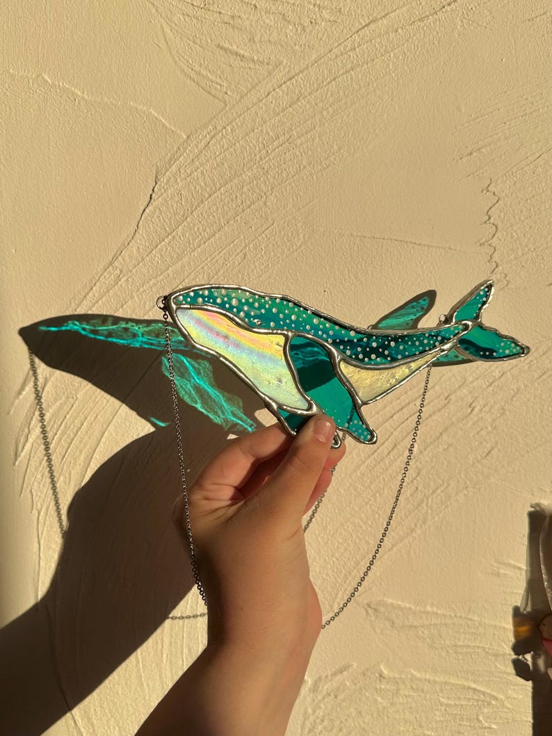 May include: A stained glass whale hanging from a silver chain. The whale is teal blue with white spots and iridescent accents.