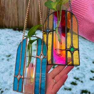 May include: Two stained glass hanging planters, each with a clear glass tube for plants. One is blue with copper detailing, the other is multi-coloured with yellow, orange, and pink. Both are suspended by chains and contain green plants.