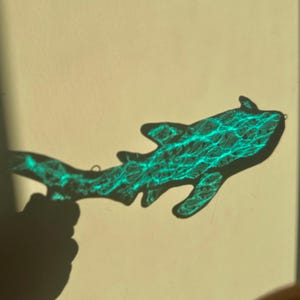 May include: A shark-shaped object with a textured, turquoise pattern is held in front of a light-colored background. The object's shadow is cast on the background, and a hand is visible holding the object.