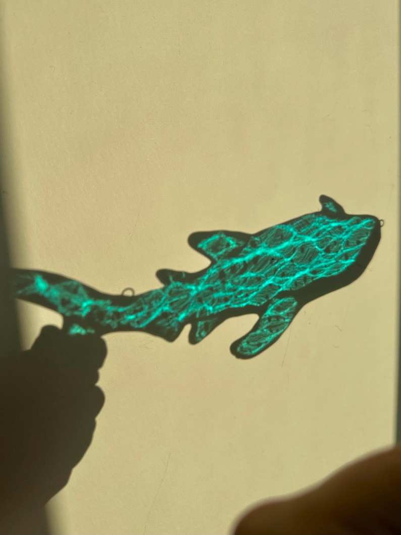 May include: A shark-shaped object with a textured, turquoise pattern is held in front of a light-colored background. The object's shadow is cast on the background, and a hand is visible holding the object.