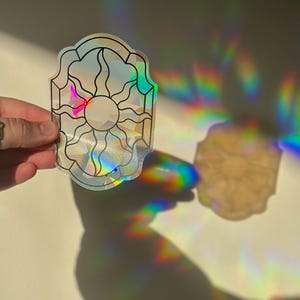 May include: A holographic sticker featuring a sun design. The sticker has a black outline and a clear centre, with rainbow colours reflecting light. The sticker is held up against a white background, with a shadow of the sticker visible.