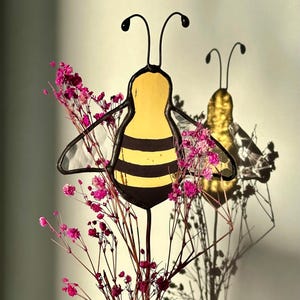 Peut inclure: Décoration d'abeille en vitrail avec des rayures jaunes et noires, des contours noirs et de délicates antennes. Des fleurs roses séchées sont devant l'abeille. L'ombre de l'abeille est visible sur le mur.
