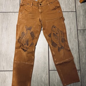 May include: Brown work pants with a straight fit and black fish designs on the knees. The pants have a label that says "Straight Fit".