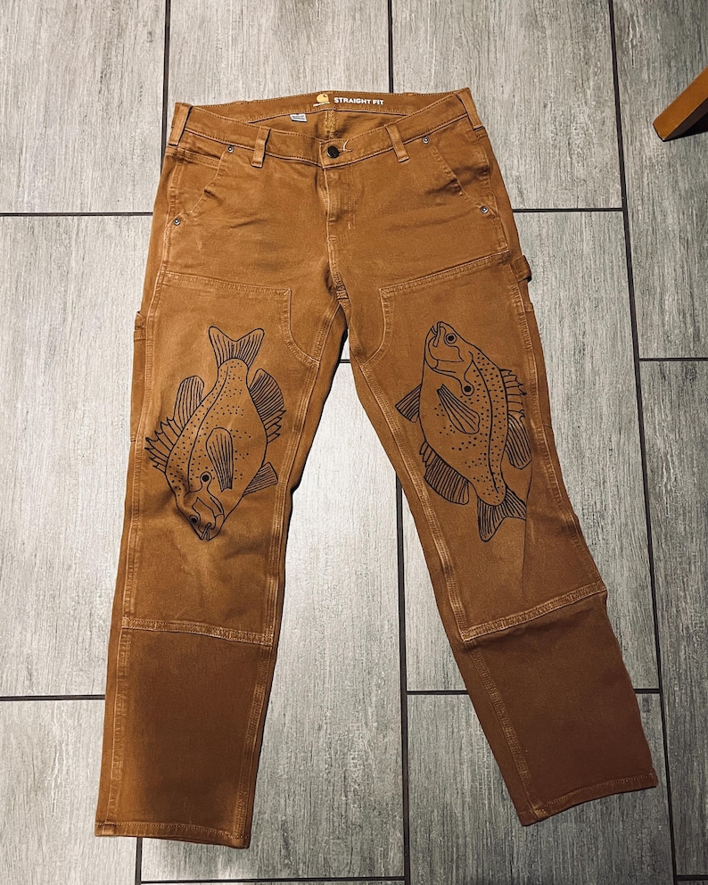 May include: Brown work pants with a straight fit and black fish designs on the knees. The pants have a label that says "Straight Fit".