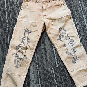 May include: A pair of tan work pants with black fish illustrations on each leg. The pants have a distressed look and are made of a durable fabric.