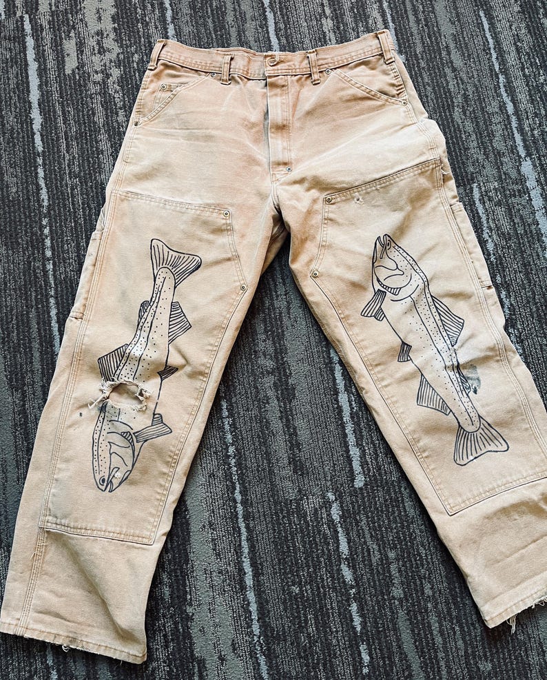May include: A pair of tan work pants with black fish illustrations on each leg. The pants have a distressed look and are made of a durable fabric.