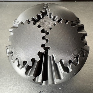 May include: A black, 3D-printed sphere with integrated gears. The sphere has a textured surface and is composed of multiple interlocking gear components. The gears are arranged around the sphere's surface, creating a complex mechanical design.