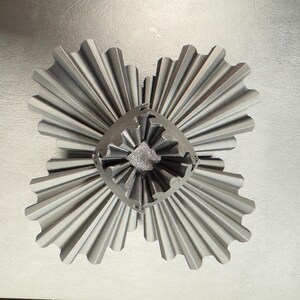 May include: A gray, metallic, four-part decorative object with a central, textured core. Each section has a fan-like design with ridged edges, creating a symmetrical, starburst appearance. The object is set against a light gray background.
