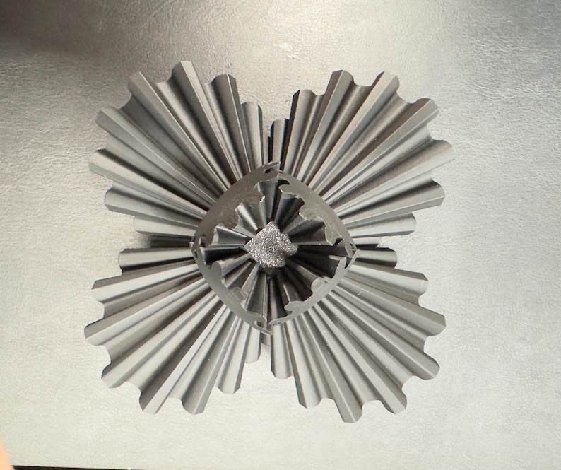 May include: A gray, metallic, four-part decorative object with a central, textured core. Each section has a fan-like design with ridged edges, creating a symmetrical, starburst appearance. The object is set against a light gray background.