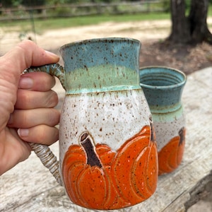 May include: Two ceramic mugs featuring a speckled white base and a blue-green rim. The lower section of each mug is decorated with embossed, bright orange pumpkins with dark brown stems. Each mug has a textured, ribbed handle.