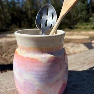May include: A ceramic utensil holder with a glossy glaze featuring blended pastel pink, purple, and cream colors. Inside the holder are a silver metal slotted spoon and a light-colored wooden spoon, both standing upright.
