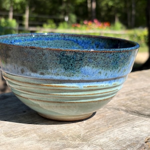 May include: A handmade ceramic bowl with a glossy finish. The bowl features a gradient of colors, transitioning from a deep blue at the rim to teal and light green towards the base. Horizontal ridges add texture to the exterior.