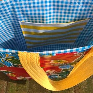 Puede incluir: Un bolso tote colorido con un estampado floral sobre un fondo rojo. El interior presenta un patr&oacute;n de cuadros azules y blancos y un bolsillo a rayas amarillas y blancas. Las correas y los ribetes amarillos completan el dise&ntilde;o.