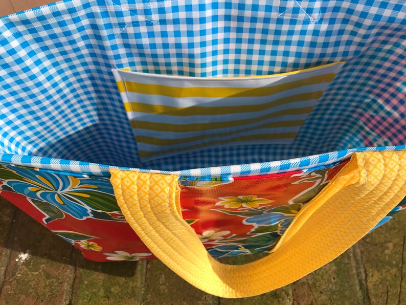 Puede incluir: Un bolso tote colorido con un estampado floral sobre un fondo rojo. El interior presenta un patr&oacute;n de cuadros azules y blancos y un bolsillo a rayas amarillas y blancas. Las correas y los ribetes amarillos completan el dise&ntilde;o.