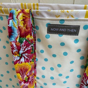 May include: Cream-colored tote bag with blue polka dots and floral handles. The handles feature a vibrant mix of red, yellow, and pink flowers. The bag has a label that reads "NOW AND THEN".