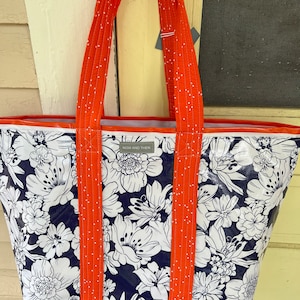 May include: A large, navy blue tote bag with a white floral print. The bag has orange straps with white polka dots and a white label that says "NOW AND THEN".