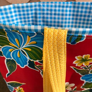 Puede incluir: Primer plano de un bolso tote con un estampado floral rojo y correas amarillas. El forro interior es de cuadros azules y blancos. El bolso presenta un dise&ntilde;o floral vibrante con flores azules y amarillas.