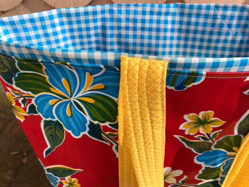 Puede incluir: Primer plano de un bolso tote con un estampado floral rojo y correas amarillas. El forro interior es de cuadros azules y blancos. El bolso presenta un dise&ntilde;o floral vibrante con flores azules y amarillas.