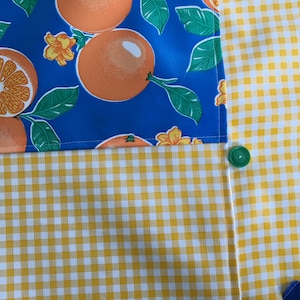 Puede incluir: Una tela azul y amarilla con un patrón de cuadros. La tela tiene un diseño de naranjas y flores en la mitad superior. Un botón verde está cosido en el lado derecho de la tela.