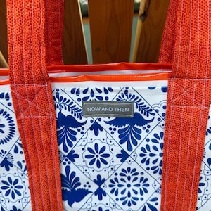 May include: A tote bag featuring a white and blue tile-inspired floral pattern with orange fabric straps. A small grey rectangular label is stitched to the front center, displaying the white text "NOW AND THEN" in a clean, sans-serif font.