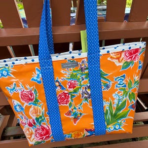 May include: Orange tote bag with a floral and butterfly pattern. The bag has blue polka dot handles and trim. The bag is labeled "NOW AND THEN".