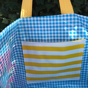 Puede incluir: Un bolso tote a cuadros azules y blancos con un bolsillo a rayas amarillas y blancas. El bolso tiene asas amarillas estampadas. El interior del bolso es visible y el fondo es una planta verde.