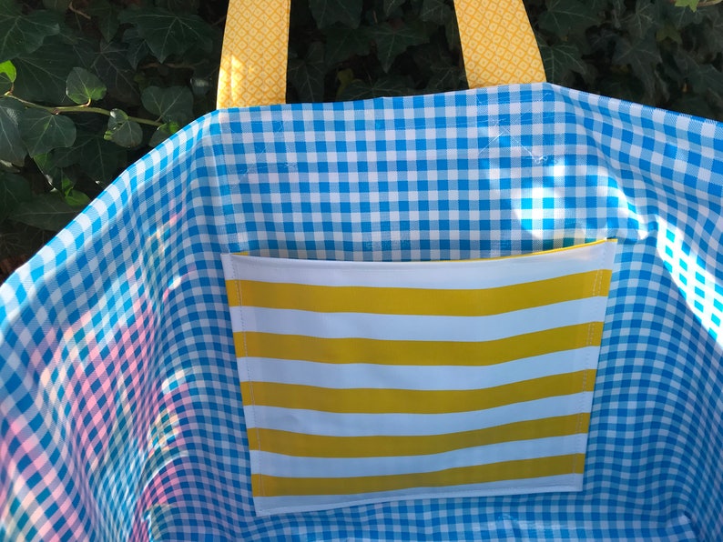 Puede incluir: Un bolso tote a cuadros azules y blancos con un bolsillo a rayas amarillas y blancas. El bolso tiene asas amarillas estampadas. El interior del bolso es visible y el fondo es una planta verde.