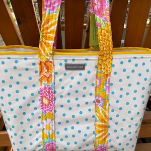 May include: A white tote bag featuring a light blue polka dot pattern and a yellow top trim. The bag has two quilted shoulder straps with a vibrant floral print in shades of pink, orange, and yellow. A grey label reads NOW AND THEN.