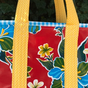 Puede incluir: Primer plano de un bolso tote con un estampado floral. El bolso tiene un fondo rojo con flores azules y blancas y hojas verdes. Las correas amarillas y un ribete de cuadros azules y blancos completan el dise&ntilde;o.