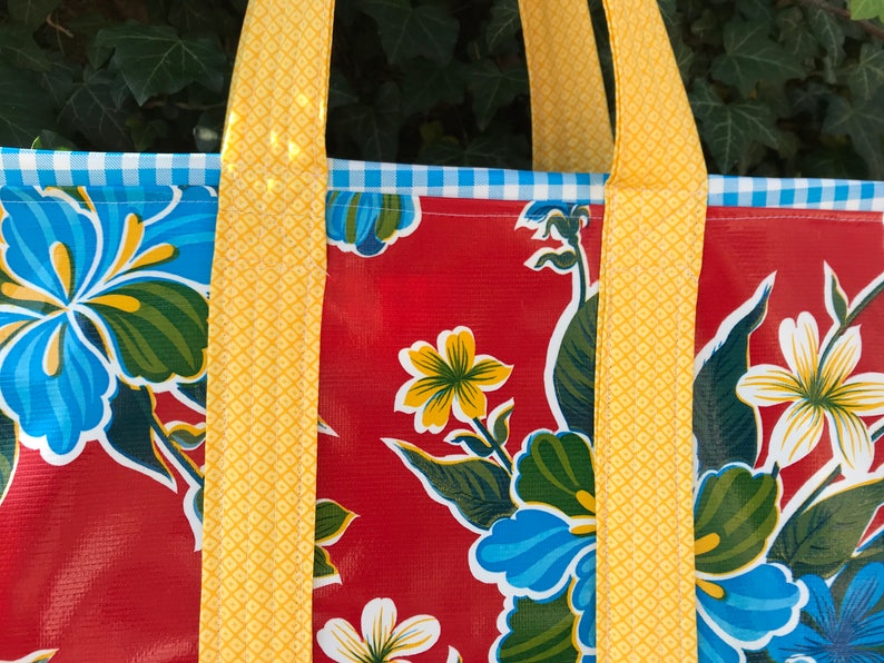 Puede incluir: Primer plano de un bolso tote con un estampado floral. El bolso tiene un fondo rojo con flores azules y blancas y hojas verdes. Las correas amarillas y un ribete de cuadros azules y blancos completan el dise&ntilde;o.