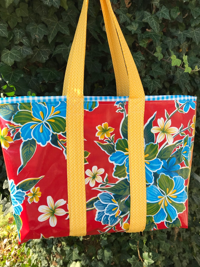 Puede incluir: Un bolso tote grande con un vibrante estampado floral. El bolso presenta un fondo rojo con flores azules y blancas y hojas verdes. Las correas amarillas y un ribete a cuadros azul y blanco completan el dise&ntilde;o. El bolso est&aacute; dise&ntilde;ado para transportar art&iacute;culos.