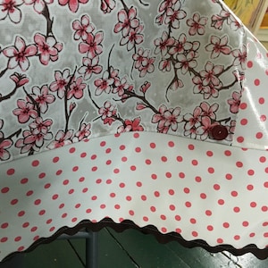 May include: A white tablecloth with a pink cherry blossom print on the top half and a white background with pink polka dots on the bottom half. The tablecloth has a brown scalloped edge.