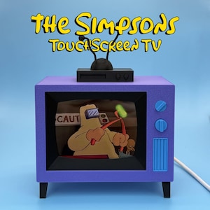 May include: A purple toy television with the words "The Simpsons Touchscreen TV" in yellow. The TV has a black antenna, blue buttons, and a screen displaying a cartoon scene. The TV is on a blue background.