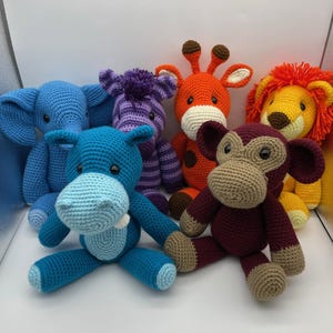 May include: Six crocheted stuffed animals, including a blue elephant, purple zebra, orange giraffe, brown monkey, blue hippopotamus, and yellow lion. The animals are all sitting on a white surface.