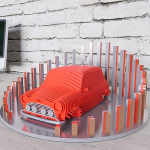 May include: A red, layered wooden car model sits on a silver circular base, surrounded by vertical wooden bars. The car model has a detailed front with headlights and a grill. The background includes a computer and a white brick wall.