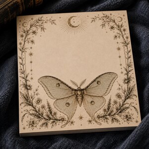 May include: A square notepad with light brown paper featuring a dark brown ink illustration of a large moth centered at the bottom. The design includes a floral border with stars and a crescent moon at the top, creating a decorative frame.