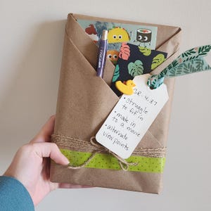 Könnte beinhalten: Ein in braunes Papier verpacktes Paket, mit Bindfaden und grünem Klebeband verschnürt. Es enthält einen Stift, Aufkleber und ein Etikett mit handschriftlichem Text: "Struggle to fit in", "made in to a movie" und "alternate view points."