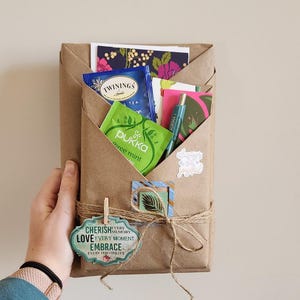 May include: A brown paper package wrapped with twine and decorated with a green leaf, a small wooden peg, and a sticker that says "Cherish every memory, love every moment, embrace every possibility". The package contains tea bags, cards, and a pen.