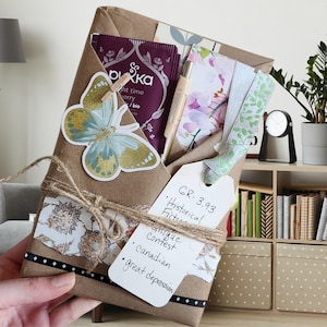 May include: A brown paper package wrapped with twine and decorated with a butterfly sticker, a floral patterned paper, and a tag with the text "GR-3.93 • Historical Fiction • Unique Contest • Canadian • Great Depression".