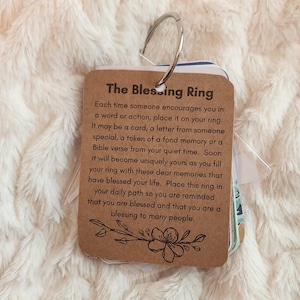 Puede incluir: Un "Blessing Ring" con un anillo de metal que sostiene varias tarjetas pequeñas. La tarjeta principal es marrón con el texto "The Blessing Ring" y un diseño floral. Las tarjetas están sobre una superficie blanca y esponjosa.
